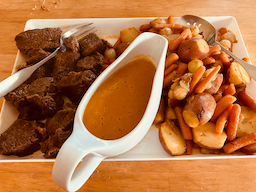 A white rectangular platter with chunks of braised beef on the left, a white gravy boat with an ochre sauce inside in the middle, and heap of braised baby carrots, baby red potatoes, and baby onions on the right