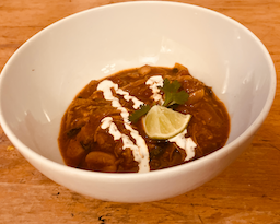 A bowl of mahogony stew with shredded pork, chopped greens, and white beans topped with a drizzle of fresh crema, a sprig of cilantro, and a lime wedge