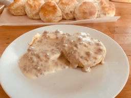 A split biscuit topped with flowing white gravy full of small sausage bits, with a sheet of golden biscuits in the background